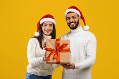 Noel Baba şapkası takan gülümseyen bir çift ellerinde kırmızı kurdeleli bir hediyeyle birlikte parlak sarı bir arka planın önünde duruyorlar. Neşeli ifadeleri bayram ruhunu yansıtıyor..