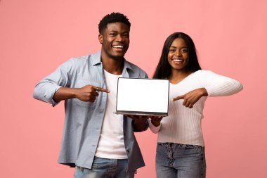 Neşeli genç siyah bir çift boş ekran ile modern bir dizüstü bilgisayar sergiliyor, bu da yararlı bir yazılım veya web sitesi öneriyor. Birlikte canlı pembe bir stüdyoda pozitiflik yayarak poz veriyorlar..