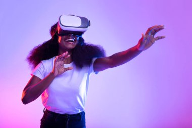 Mutlu genç siyah bir bayan sanal gerçeklik gözlüğü takıyor ve hayali bir ekrana dokunmak için uzanıyor. Canlı neon ışıklarla çevrili büyüleyici bir sanal gerçeklik oyununa daldı..