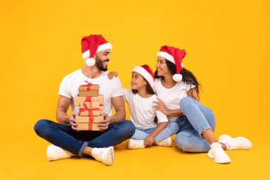 Neşeli bir aile yerde oturuyor, Noel Baba şapkaları takıyor, gülümsüyor ve gülüyor. Önlerinde hediyeleri sarıp neşeli tatil ruhlarını vurguluyorlar..