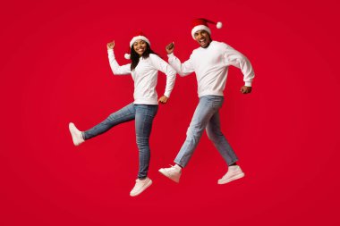 Eşleşen kış şapkaları ve sıradan kazaklar giyen genç bir çift parlak kırmızı bir arka planda mutlu bir şekilde dans ediyor. Birlikte bayram ruhunun tadını çıkarırlarken neşeli ifadeleri neşeli bir neşe yansıtır..