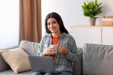 Güzel, genç bir Arap kadın evinde rahat bir şekilde oturuyor, bilgisayarını internette çalışmak için kullanıyor. Bir Webinar izlerken bir fincan kahveden zevk alıyor, rahat bir atmosfer yaratıyor..