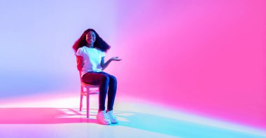 Neşeli siyah bir kadın beyaz bir sandalyede oturuyor, izleyicileri boş neon ışıklı arka planlarını reklamları için kullanmaya davet ediyor. Neşeli ifadesi sahneye sıcaklık katıyor..