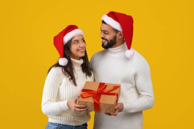 Noel Baba şapkası takan bir adam ve kadın birbirlerine gülümserken güzel bir hediye tutuyorlar. Sahnede neşeli sarı bir arka plan var, tatil ruhu için mükemmel..