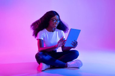 Milenyum kadını yerde otururken tablet kullanıyor. Çevrimiçi bir konferansa katılırken gülümsüyor. Neon ışıkları, uzak çalışmaları için canlı bir atmosfer yaratıyor..