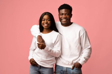 Güzel bir Afro-Amerikan çifti benzer kıyafetler içinde gülümseyerek ve el ele tutuşarak bir arada duruyor. Pembe bir stüdyo ortamında birbirlerini kucaklarken sevgi ve neşe yayıyorlar..