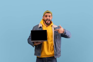 Bir adam bir elinde dizüstü bilgisayarını tutarken geniş bir şekilde gülümsüyor ve diğer eliyle onu işaret ediyor. Kot ceketinin altına sarı bir kapüşonlu giyiyor, mavi arka planda..