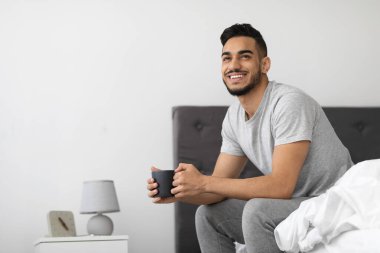 Genç bir Arap adam bir fincan kahveyle yatağında oturur, gülümser ve rahat yatak odasında huzurlu bir sabah geçirir. Ayar güne olumlu ve rahat bir şekilde başlamasını yansıtıyor..