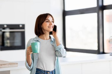 Genç bir kadın modern bir mutfakta elinde bir deniz mavisi kupasıyla telefonuyla sohbet ediyor. Sabahın keyfini çıkarırken güneş ışığı pencerelerden akar..