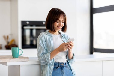 Genç bir kadın mutfak tezgahına yaslanırken akıllı telefonunu kontrol ederken gülümsüyor. Tezgahın üzerinde duran bir kahve fincanı, parlak uzayda rahat bir atmosfer yaratıyor..