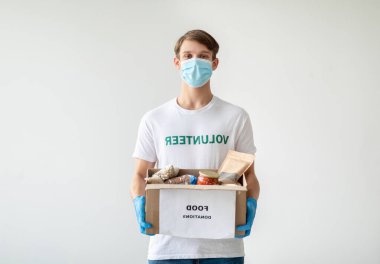 Gönüllü tişörtlü, sağlık maskeli ve eldivenli genç bir adam ışık duvarına yaslanıyor. Elinde yiyecek dolu bir bağış kutusu var ve kameraya gülümseyerek bakıyor..