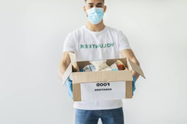 Gönüllü tişörtü ve tıbbi maskesi olan bin yıllık Arap bir adam konserve yiyecek ve paketlerle dolu bir kutu gösteriyor. Halkın desteğini vurgulayarak hafif bir stüdyonun önünde duruyor..