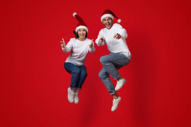 Neşeli bir çift Noel şapkaları giyerken havaya zıplıyor. Günlük kıyafetler giyip neşeli ifadeler sergiliyorlar. Bayram ruhunu kutluyorlar..