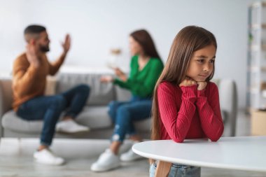 Hafifçe aydınlatılmış bir oturma odasında, ailesi arka planda kavga ederken genç bir kız üzgün görünür. Yalnız ve ihmal edilmiş hisseden çocuk, anne ve babası ayrılmaları hakkında tartışırken ileriye bakar..