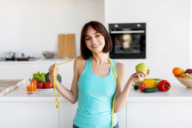 Neşeli bir kadın parlak bir mutfakta, bir elinde bir elma, diğer elinde bir mezura ile duruyor. Taze meyve ve sebzeler etrafını sarmış, sağlıklı bir yaşam tarzını teşvik ediyor..