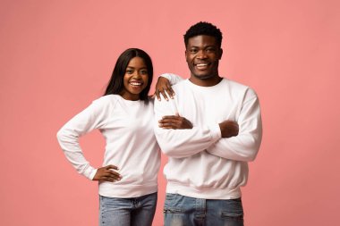 Neşeli genç siyah bir çift pembe bir stüdyo arka planında sarılıyor. Kadının uzun saçları ve erkek arkadaşına sarılırken gülümsemesi var. İkisi de aynı günlük kıyafetleri giyiyor..