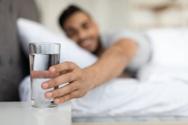 Genç bir Arap sabahleyin komodindeki bir bardak suya uzanırken yatakta dinlenir. Sahne, güne doğal ışıkla sakin bir başlangıç gösteriyor..