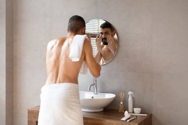 Genç bir Arap adam, göz altı bölgesine krem sürerken banyo aynasında yansımasına bakar. Kendine güveni tam görünüyor ve yüz bakımına odaklanmış, havluya sarılmış..