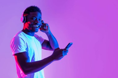 Neşeli genç siyah bir adam kablosuz kulaklık takıyor ve akıllı telefonunu kullanıyor. Etrafı parlak neon ışıklarıyla çevrili ve müzikten ya da sesli kitaptan hoşlanıyor gibi görünüyor..