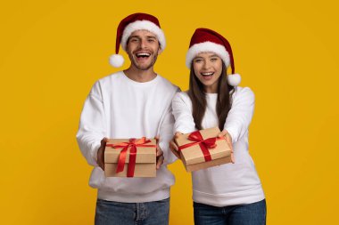 Noel Baba şapkası takan iki neşeli kişi parlak sarı bir arka planda güzel paketlenmiş Noel hediyeleri taşıyorlar. Gülümsüyorlar, bayram neşesi yayıyorlar..