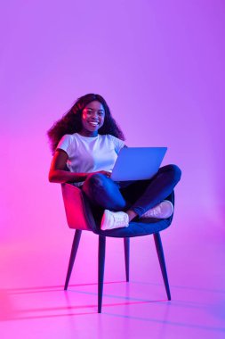 Genç siyahi kadın dizüstü bilgisayarını kullanırken sırıtarak koltukta oturuyor. Oda canlı neon ışıklarıyla aydınlatılıyor, canlı bir atmosfer yaratıyor..
