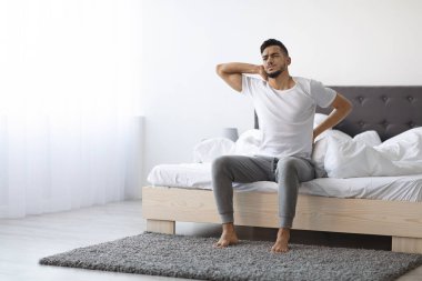 Genç bir Orta Doğulu rahatsız bir yatağın kenarında oturur, kaşlarını çatar ve sırtına ve boynuna masaj yapar. Sabahın erken saatlerinde basit bir yatak odasında acı belirtileri gösteriyor..