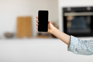 Bir insan rahat bir mutfakta kameraya doğru akıllı bir telefon tutar. Ayarda tarafsız renkler ve sıcak bir ortam yer alıyor, bu da iletişim veya paylaşım için rahatlatılmış bir atmosfer izlenimi veriyor..