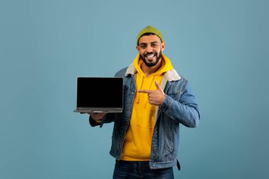 Genç bir adam dizüstü bilgisayarıyla güvenle ayakta duruyor, gülümsüyor ve ekranı işaret ediyor. Kot ceketinin altına yeşil bir bere ve sarı bir kapüşon giyiyor..