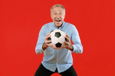 Gri saçlı bir adam elinde siyah beyaz bir futbol topu tutarken geniş bir gülümseme sergiliyor. Kareli bir gömlek giyiyor ve parlak kırmızı bir arkaplanın önüne yerleştirilmiş..