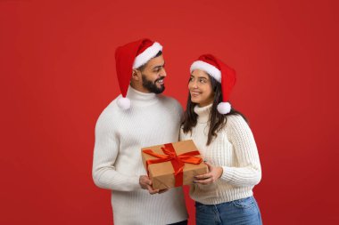 Neşeli Arap çift canlı kırmızı bir zemin üzerinde duruyor, birbirlerine gülümserken güzel paketlenmiş bir Noel hediyesini tutuyorlar..