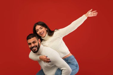 Sevgi dolu bir Arap, Orta Doğulu kız arkadaşını sırtında taşıyor. Hem gülümsüyor hem de canlı kırmızı stüdyo arkaplanının karşısında anın tadını çıkarıyor..