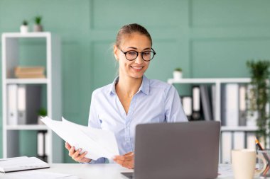 Kadın girişimci iş raporlarını tutarken dizüstü bilgisayarına odaklanıyor. Modern bir ofiste şık bir masada oturuyor, organize dosyalar ve hoş geldin atmosferiyle çevrili..