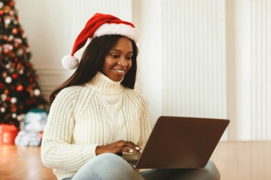 Noel Baba şapkalı bir kadın dizüstü bilgisayar kullanarak bacak bacak üstüne atmış şekilde yerde oturuyor. Çalışırken gülümsüyor, arka planda süslü bir Noel ağacı ve yakında da tatil hediyeleri var..