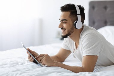 Genç, mutlu bir Arap yatakta kablosuz kulaklık takıyor. İnternette gezinmek için dijital tablet kullanıyor. Rahat ve rahat yatak odasında..