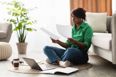 Parlak bir yaşam alanında, bir kadın yerde oturuyor, etrafı kağıtlar ve dizüstü bilgisayarlarla çevrili. Belgelerine odaklanmış, günlük giysiler giyiyor ve huzurlu bir çalışma seansının tadını çıkarıyor..