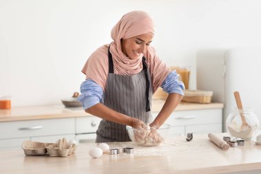 Başörtüsü takan bir kadın, temiz bir mutfakta hamuru zevkle yoğuruyor. Güneş ışığı alan alan, pişirmeye hazırlanırken sıcak bir atmosferi yansıtıyor..