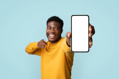 Sarı elbiseli neşeli Afro-Amerikan genç adam mavi stüdyo arka planında poz verirken boş beyaz ekranlı modern cep telefonunu gösteriyor, en yeni mobil uygulamayı öneriyor.