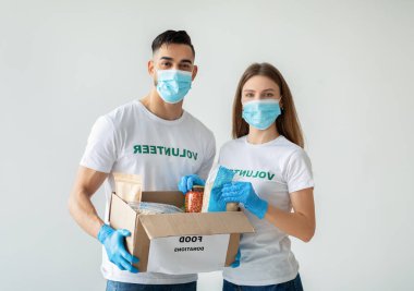 İki mutlu melez ırk gönüllüsü, yiyecek bağışlarını bir kutuya koyarken tıbbi maske ve mavi eldiven takıyor..