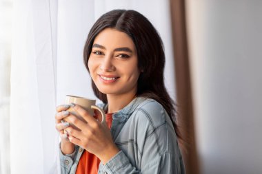 Genç bir Arap kadın pencerenin kenarında oturur, aromatik kahveyi yudumlar ve huzurlu bir sabah geçirir. Sıcak bir şekilde gülümsüyor, evinin rahatlığı ve içkisinin verdiği basit zevkin tadını çıkarıyor..