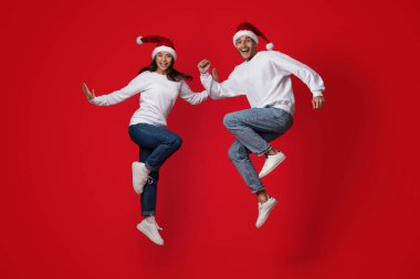 Beyaz süveterler ve Noel Baba şapkaları giyen neşeli bir çift havada zıplıyor. Canlı kırmızı bir arkaplan önünde mutluluk ve heyecanı ifade ederler. Bayram kutlamaları için mükemmeldirler..