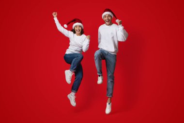 İki arkadaş coşkuyla zıplıyorlar, Noel Baba şapkaları ve günlük giysiler giyiyorlar. Neşeli ifadeleri bayram ruhunu canlı kırmızı bir ortamda yansıtıyor. Kutlamalar için ideal..