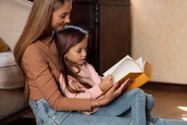 Bir anne kızıyla rahat bir oturma odasında oturup açık bir kitaptan birlikte kitap okur. Sıcak atmosfer, öğleden sonra güneşinde birbirlerine bağlanma deneyimlerini arttırır..