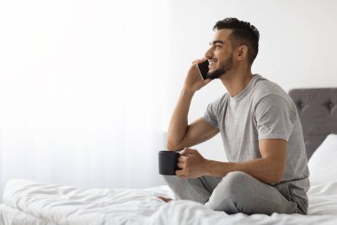 Mutlu genç bir Arap, cep telefonuyla konuşurken elinde bir fincan kahveyle yatakta rahatça oturur. Hoş bir sohbetten zevk alırken gülümsüyor, yatak odasında rahat bir atmosfer yaratıyor..