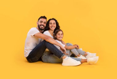 Üç kişilik mutlu bir aile, canlı sarı bir arka planda birlikte oturur, gülümser ve eğlenirler. Günlük beyaz gömlekler ve kot pantolon giyiyorlar, sıcaklık ve neşe saçıyorlar..