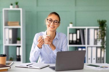 Neşeli bir ofis çalışanı masasında oturuyor, gülümsüyor ve elinde bir fincan kahve tutuyor. Aktif olarak bilgisayarında çalışıyor. Bitki ve dosyalarla dolu, modern bir ofiste..