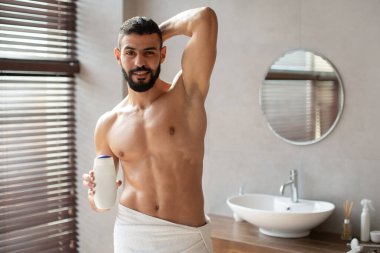 Yakışıklı genç adam şık bir banyoda havluya sarılı duruyor. Elinde bir şampuan şişesi tutuyor, doğal ışıkla çevrelenmişken kendinden emin bir poz veriyor..