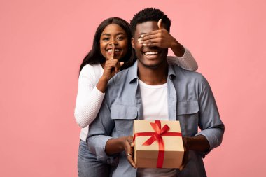 Genç, güzel, siyah bir kadın, erkeğin gözlerini kapatıyor. Elinde bir hediye kutusu tutarken heyecanını dile getiriyor..
