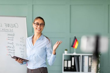 Neşeli bir bayan öğretmen çalışma alanından online Almanca dersi veriyor. Covid-19 salgını sırasında uzak öğrencilere dil bilgisi kavramlarını açıklamak için akıllı telefonunu kullanıyor..