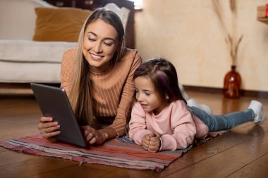 Bir anne kızının yanında yerde yatıyor, her ikisi de tabletle meşgul. Gülüşlerini paylaşıyorlar, davet edilen oturma odalarında sıcak bir atmosfer yaratıyorlar..