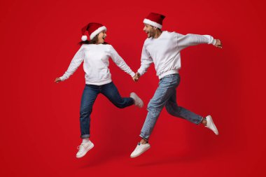 İki neşeli birey havaya atlarken el ele tutuşuyorlar. Parlak kırmızı Noel Baba şapkaları ve rahat kazaklar giyip Noel ruhunu saf mutlulukla kutluyorlar..